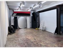 Fotostudio mit professioneller Ausrüstung, darunter Lichtreflektoren, Softboxen, eine weiße Hintergrundwand, eine Leiter und ein Ventilator. Der Raum hat eine rohe Betonbodenoptik und eine Decke mit sichtbaren Streben sowie schlichte weiße Vorhänge an der Seite.