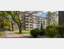 Wohnblock mit mehreren Etagen und Balkons in hellen Beige- und Grautönen, umgeben von Grünflächen, Bäumen und Büschen; ein gepflasterter Weg führt durch die parkartige Umgebung.