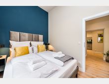 Modernes Schlafzimmer mit Doppelbett, gepolstertem grauen Kopfteil und blau gestrichener Akzentwand; weiße Bettwäsche mit gelben Kissen und Handtüchern auf dem Bett. Rechts führt eine offene Tür in einen Flur mit Holzboden, von dem aus eine Küche zu sehen ist.