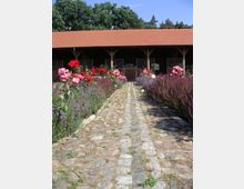 Pflasterweg, gesäumt von blühenden Beeten mit roten, rosa und violetten Blumen, führt zu einem langgezogenen Stallgebäude mit roten Ziegeln und Holzstützen, im Hintergrund von Bäumen umgeben.