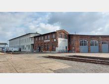 Backsteingebäude eines Bahnbetriebswerks mit der Beschriftung „Lokdienstleitung“ und „Abteilung Tb“, daneben ein weiß verputztes Bürogebäude. Im Vordergrund befinden sich Bahngleise sowie ein befestigter Vorplatz.