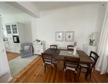 Esszimmer in einem hellen Wohnraum mit Holzdielenboden. Zu sehen sind ein dunkler Holztisch mit vier Stühlen, eine weiße Kommode, zwei gerahmte Bilder an der Wand, ein Sessel in grauem Stoff und eine weiße Einbauküche im Hintergrund. Ein seitlicher Fensterbereich mit einem transparenten Vorhang bringt Tageslicht herein.