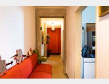 Ein schmaler Flur mit beigen Wänden, einem roten Sofa links und einer kleinen grünen Sitzgelegenheit. Am Ende des Flurs hängt ein roter Vorhang, und an den Wänden befinden sich dekorative Bilderrahmen; rechts ist ein Durchgang zu einer Küche mit Geschirr und einer Kaffeemaschine.