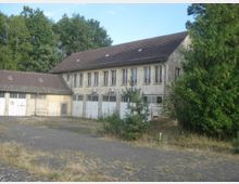 Ein zweistöckiges, verwittertes Gebäude mit Satteldach und mehreren rechteckigen Fenstern, umgeben von großen Bäumen. Im Vordergrund ist ein gepflasterter, teils überwucherter Hofbereich zu sehen, daneben ein kleiner Baum und Büsche.