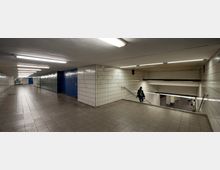 Ein unterirdischer U-Bahn-Zugang mit weiß gefliesten Wänden, einfachem Beleuchtungssystem an der Decke und Treppen, die zu den Bahnsteigen führen. Ein langer Flur mit blauen Türen und Werbetafeln am Ende ist zu sehen.