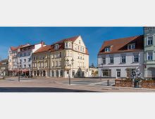 Städtischer Platz mit historischen Wohn- und Geschäftshäusern im Hintergrund, die helle Fassaden und rote Ziegeldächer haben. Im Vordergrund befinden sich eine gepflasterte Fläche, Bänke, Laternen und ein moderner Kunstbrunnen.