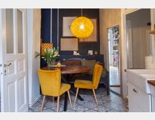 Gemütliche Essecke in einer Küche mit rundem Holztisch, zwei gelben Polsterstühlen und einer eingebauten Sitzbank vor einer dunkelblauen Akzentwand mit gerahmten Bildern. Über dem Tisch hängt eine große runde Pendelleuchte, und der Boden ist mit gemusterten Fliesen ausgelegt; rechts ist ein weißes Spülbecken und ein Durchgang zu einem Flur zu sehen.