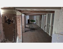 Verlassener Innenflur eines alten Gebäudes mit abblätternder Farbe, Stützpfeilern und freiliegenden Deckenbalken. Rechts verläuft eine Reihe hoher Fenster- oder Türrahmen, an Wänden und Türen sind Graffiti und Risse im Putz zu sehen.