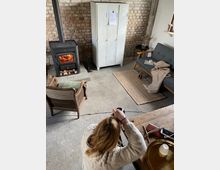 Ein gemütlich eingerichteter Raum mit freiliegenden Ziegelwänden, einem schwarzen Kaminofen mit Holzlager, einem weißen Metallschrank und verschiedenen Sitzecken, darunter ein graues Sofa mit einer Decke. Im Vordergrund sitzt eine Person an einem Holztisch, während im Hintergrund Teppiche und kleine Tische den industriellen Stil ergänzen.