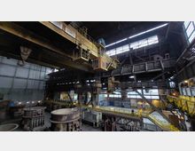 Innenansicht einer Industriehalle mit hohen Decken, einem großen Kran an der Deckenkonstruktion und Metallplattformen mit Geländern. Im Vordergrund befinden sich massive Gussformen und Maschinen, während im Hintergrund große Fenster für Licht sorgen.