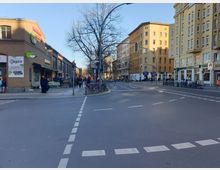 Straßenecke in einer Stadt mit mehrstöckigen Wohn- und Geschäftsgebäuden aus gelbem und rotem Mauerwerk. Im Vordergrund ein Zebrastreifen, im Hintergrund Fahrradständer, ein Supermarkt („Veganz“) und Fußgänger bei sonnigem Wetter.