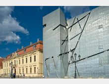 Historisches Gebäude mit gelber Fassade und rotem Dach neben einem modernen, silbernen Bau mit futuristischer Metallverkleidung und unregelmäßigen Linien; im Vordergrund eine Straßenlaterne und Passanten. Der Himmel ist blau mit vereinzelten Wolken.
