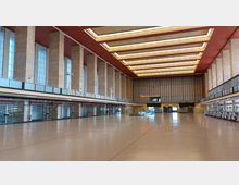 Weitläufiger, historischer Terminalbereich eines Flughafens mit hohen Säulen, großen Fenstern und einer Deckenbeleuchtung in geometrischem Design. Im Hintergrund befinden sich eine Uhr, ein Leuchtschriftzug "Restaurant" und mehrere geschlossene Schalter.