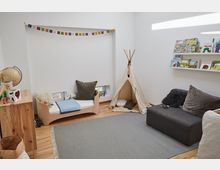 Helles Kinderzimmer mit Holzboden, einem kleinen Zelt mit Kissen, einem Kinderbett, einem grauen Teppich, einem Sofa und einem Bücherregal an der Wand. Eine bunte Wimpelkette hängt an der Wand, und auf einer Holzkommode steht ein Globus.