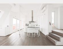Moderne, helle Wohnung mit offenem Küchenbereich, weißen Wänden und dunklem Holzboden. Der Raum verfügt über große Fenster, eine Kochinsel mit zwei weißen Stühlen und eine Treppe, die zu einem höher gelegenen Bereich führt.