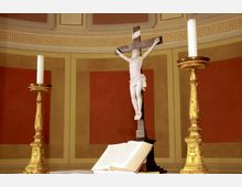 Kreuzdarstellung in einer Kirche mit einer weißen Jesusfigur auf einem Holzkreuz, flankiert von zwei goldenen Kerzenleuchtern mit weißen Kerzen. Im Vordergrund liegt ein aufgeschlagenes Buch auf einem Altar, im Hintergrund befindet sich eine verzierte Wand mit geometrischen Mustern.