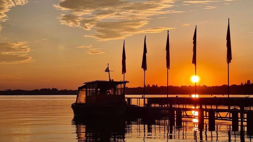 Sonnenuntergang an einem ruhigen See mit einem Bootsanleger, auf dem ein Boot und mehrere Fahnenmast-Silhouetten stehen. Im Hintergrund reflektiert die tief stehende Sonne orangefarbenes Licht auf der Wasseroberfläche.