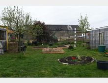 Eine Außenaufnahme eines Gartens mit mehreren Hochbeeten, jungen Bäumen und einer Wiese. Im Hintergrund sind ein Gewächshaus, ein landwirtschaftliches Gebäude und ein Traktor zu sehen.