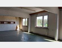 Ein leerer, renovierungsbedürftiger Raum mit zwei großen Fenstern, die viel Licht hereinlassen und den Blick auf eine bewaldete Landschaft bieten. Die Wände zeigen Abnutzungsspuren, es gibt Heizkörper unter den Fenstern, und ein Regal ist an der linken Wand angebracht.