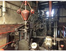 Alte industrielle Maschinenanlage in einem Fabrikraum mit Backsteinwänden. Sichtbar sind Zahnräder, Rohrleitungen, ein roter Fliehkraftregler sowie verschiedene Metallkomponenten, umgeben von abgenutzten Wänden und einer eher dunklen Umgebung.