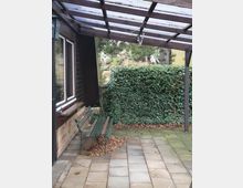 Außenbereich mit gepflastertem Boden, einer grünen Metallbank und einer Pergola mit transparentem Dach. Im Hintergrund sichtbare Efeuwand und einige Bäume.