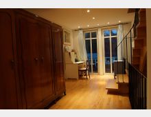 Ein Wohnraum mit Holzboden, einem großen vintage Kleiderschrank links, einem weißen Schreibtisch mit Stuhl vor bodentiefen Fenstern mit Vorhängen rechts; daneben eine Holztreppe mit Metallgeländer, die nach oben führt.