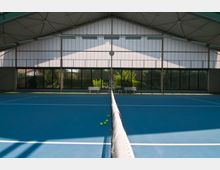 Innenansicht eines überdachten Tennisplatzes mit blauem Bodenbelag. Im Hintergrund befinden sich großflächige Fensterfronten, durch die grüner Außenbereich sichtbar ist, sowie eine Schiedsrichterleiter und zwei Bänke.