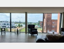 Großer Raum mit Panoramafenstern, die den Blick auf eine grüne Landschaft mit Fluss, einer modernen weißen Villa und einem historischen Backsteingebäude freigeben. Im Vordergrund stehen Ledersessel, Couchtische und Polstermöbel auf einem Holzfußboden.