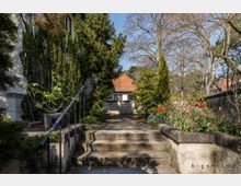 Ein kleiner Gartenweg mit Treppenstufen, umgeben von dichtem Grün, Sträuchern und bunten Tulpenbeeten. Im Hintergrund steht ein Haus mit roten Ziegeldach, eingebettet in eine baumreiche Umgebung.