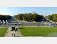 Blick auf eine große Straßenkreuzung in einer Stadt, gesäumt von Bäumen und Grünflächen. Im Vordergrund befinden sich eine Treppe und eine kleine unterirdische Öffnung, während im Hintergrund der Verkehr und umliegende Gebäude sichtbar sind.