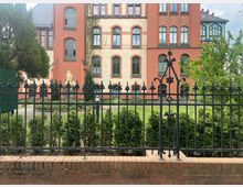 Historisches Backsteingebäude mit rot-beigen Fassaden und grünen Fensterrahmen, umgeben von einem schmiedeeisernen Zaun und gepflegter Bepflanzung; im Vordergrund steht eine Statue hinter dem Zaun.