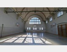 Große Sporthalle mit hoher, gewölbter Decke und großem Bogenfenster an der Stirnseite, durch das viel Tageslicht auf den markierten Hallenboden fällt. An den Wänden sind Basketballkörbe, Turngeräte und mehrere hohe Fenster angebracht.