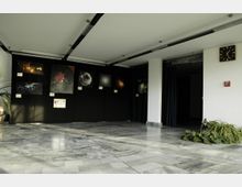 Ausstellungshalle mit Marmorboden und einer schwarzen Wand, an der Bilder von Galaxien und Weltraumphänomenen hängen. Rechts sind Vorhänge, ein Schild mit der Aufschrift "Heliostat" und eine Topfpflanze sichtbar.