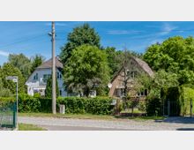 Zwei Einfamilienhäuser in einer grünen Wohngegend, umgeben von Bäumen und Hecken. Das linke Haus ist weiß, das rechte aus Backstein, beide mit Satteldächern, davor ein Gehweg und ein Straßenschild mit der Aufschrift "Schwarzwildweg".