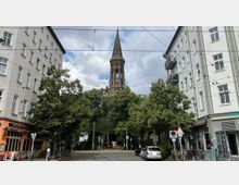 Straßenszene in einer Stadt mit Blick auf einen Kirchturm im Zentrum, flankiert von zwei mehrstöckigen Wohngebäuden. Im Vordergrund sind Bäume, Straßenschilder, Autos und ein Café mit Sitzplätzen zu sehen.