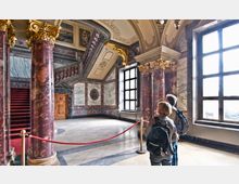 Prunkvolle Innenansicht eines historischen Gebäudes mit roten Marmorsäulen, goldenen Kapitellen und einer reich verzierten Decke. Im Raum befinden sich Besucher, die durch eine Absperrung vom mit rotem Teppich bedeckten Treppenaufgang getrennt sind; große Fenster lassen Tageslicht hinein.