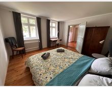 Schlafzimmer mit Holzboden, einem Doppelbett mit gemusterter Tagesdecke, zwei Fenstern mit dunklen Vorhängen, einem Schreibtisch und Stuhl sowie einem Kleiderschrank aus Holz. Eine offene Tür führt in einen Flur mit Parkettboden.