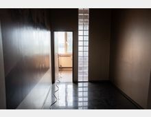 Schmaler, leerer Innenflur mit glänzendem Fliesenboden und glatten Wänden, am Ende eine offene Tür zu einem hellen Raum mit Fenster und Heizkörper. Rechts neben der Tür verläuft ein vertikales Band aus Glasbausteinen, und ein Kabel liegt über den Boden in Richtung Tür.