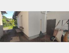 Ein kleiner gepflasterter Hof mit einem weißen Gebäude, an dessen Wand eine Gartenbank, ein Tisch und ein aufrechter Rechen lehnen. Rechts ist eine Holztür sowie eine Schubkarre, im Hintergrund sind Büsche und Bäume sichtbar.