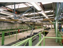 Halle eines Wartungswerks für Züge mit offenen Arbeitsgruben, grünen Geländern, metallischen Laufstegen und Stahlträgern an der Decke. Die Umgebung ist gut beleuchtet und zeigt technische Ausrüstung zwischen den Arbeitsbereichen.