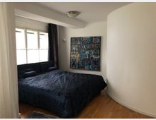 Schlafzimmer mit einem großen Bett, dunkelblauer Tagesdecke und dunkelblauen Kissen, Holzboden und einem großen Fenster mit Vorhängen. An der Wand hängt ein farbenfrohes Gemälde, und rechts ist eine gebogene, weiße Wand sichtbar.