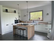 Moderne Küche mit weißer Einbauküche, Holzarbeitsplatten und einer zentralen Kücheninsel mit Gasherd. Große Fenster lassen viel Tageslicht herein, und Hocker stehen an der Insel unter drei hängenden Glühbirnenlampen.