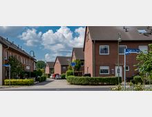 Straßenszene in einem Wohngebiet mit roten Ziegelhäusern und Satteldächern, umgeben von kleinen Gärten und Hecken. Im Vordergrund Straßenschilder mit den Beschriftungen „Paderborner Ring“ und „Gathener Weg“ sowie parkende Autos in der Nebenstraße.