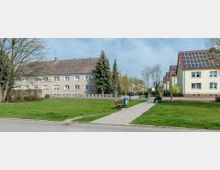 Wohnsiedlung mit mehrstöckigen Gebäuden, teils mit roten Dächern und Solarpaneelen. Im Vordergrund eine Wiese mit Bäumen, einem schmalen Gehweg und einer Sitzbank.