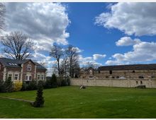 Weitläufige Grünfläche mit einer historischen Steinmauer, dahinter ein langes Gebäude aus Naturstein. Links ein zweistöckiges, rotes Backsteingebäude mit weißen Fensterrahmen und einem schwarzen Schrägdach. Im Hintergrund sind Bäume und ein bewölkter Himmel zu sehen.