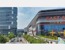 Moderne Einkaufs- und Bürogebäude in städtischer Umgebung. Links steht ein hoher Glas- und Betonbüroturm, rechts ein großes, geschwungenes Einkaufszentrum mit spiegelnder Glasfassade und dem Schriftzug „East Side“, unten Bäume und eine Straße mit parkenden Autos.