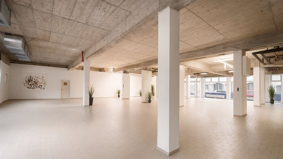 Großer, leerer Innenraum im Industrie‑Loft-Stil mit Sichtbetondecke, weißen Stützsäulen und hellem Fliesenboden. Weiße Trennwände mit einem Kunstbild, mehrere Topfpflanzen sowie große Fensterfronten zur Straße prägen den Raum.