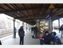 Bahnhofssteig mit überdachter Plattform und sichtbarer Bahnsteigbeschilderung "Rathaus Steglitz". Mehrere wartende Personen sind zu sehen, einige sitzen auf einer Bank, andere stehen. Im Hintergrund verlaufen Bahngleise neben einem Wohnviertel.
