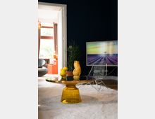 Wohnzimmer mit modernem Design: ein runder Couchtisch mit gelbem Glasfuß, darauf gelbe Keramikskulpturen, daneben ein filigraner Stuhl aus Metallgittern. Im Hintergrund eine geöffnete Tür, die zu einem helleren Raum mit großen Fenstern führt.