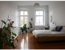 Helles Schlafzimmer mit Holzboden, einem Bett mit weißer Bettwäsche und blauen Kissen sowie einem Schreibtischbereich mit einem schwarzen Bürostuhl vor zwei großen Fenstern. Zimmerpflanzen und minimalistische Möbel ergänzen die Einrichtung.